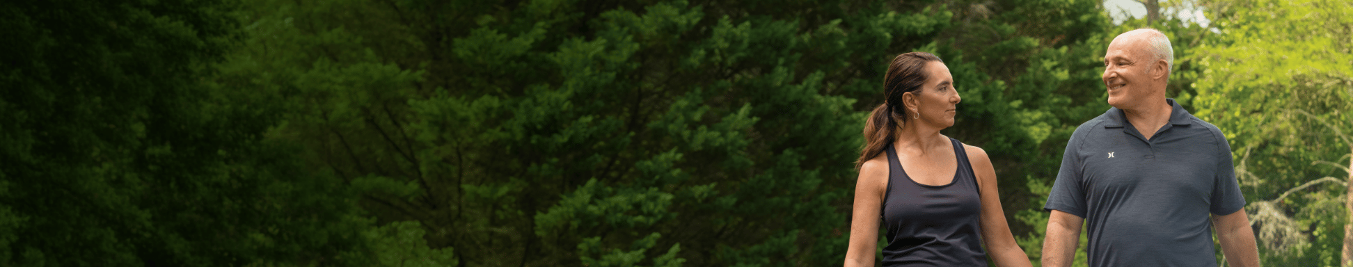 Una pareja se toma de la mano y camina por un sendero en un parque frondoso y verde, vestidos con ropa deportiva informal, transmitiendo una sensación de relajación y felicidad.
