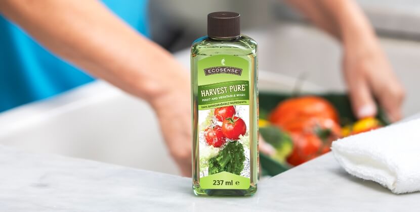 A bottle of Harvest Pure sits on a kitchen counter. Blurred background shows hands washing produce.