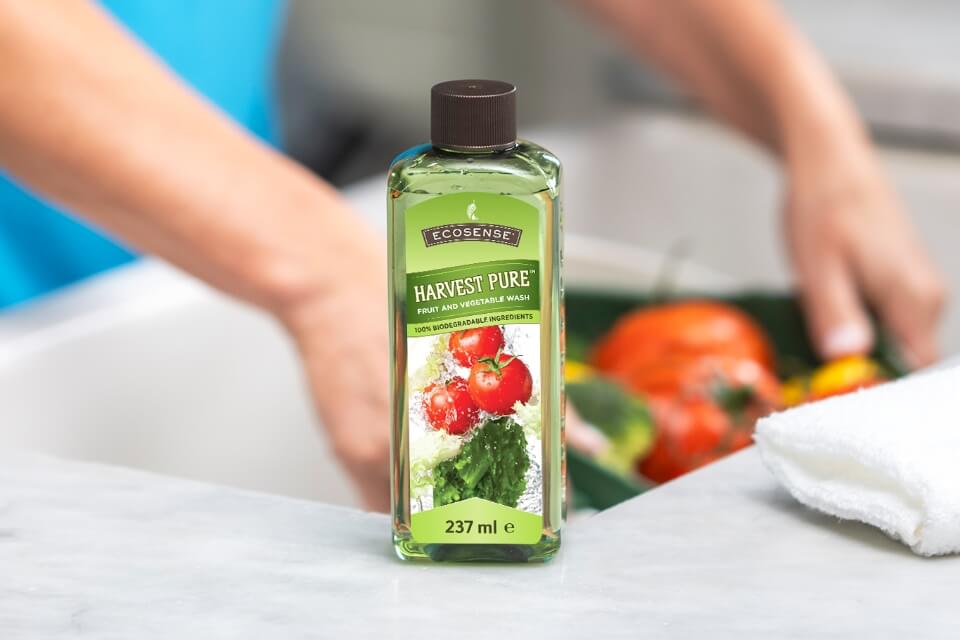 A bottle of Harvest Pure sits on a kitchen counter. Blurred background shows hands washing produce.