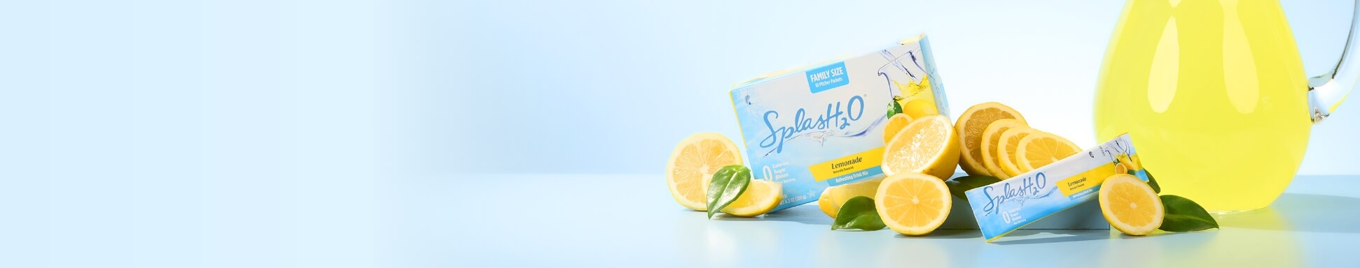 A box and packet of SplashH2O lemonade mix are surrounded by fresh lemon slices and leaves, beside a glass pitcher of lemonade, on a light blue surface.