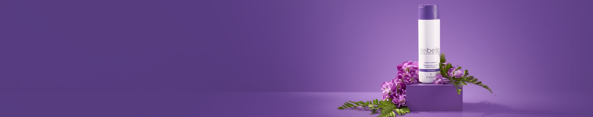 Una botella del champú morado Sei Bella sobre un bloque morado, rodeada de flores moradas y helechos verdes, con un fondo morado.