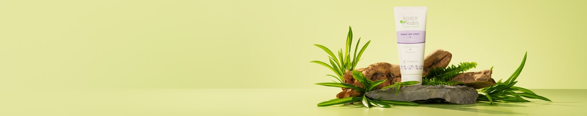 A tube of Koala Kubs Baby Diaper Rash Cream stands on a stone, surrounded by lush green leaves and driftwood, set against a soft green background.