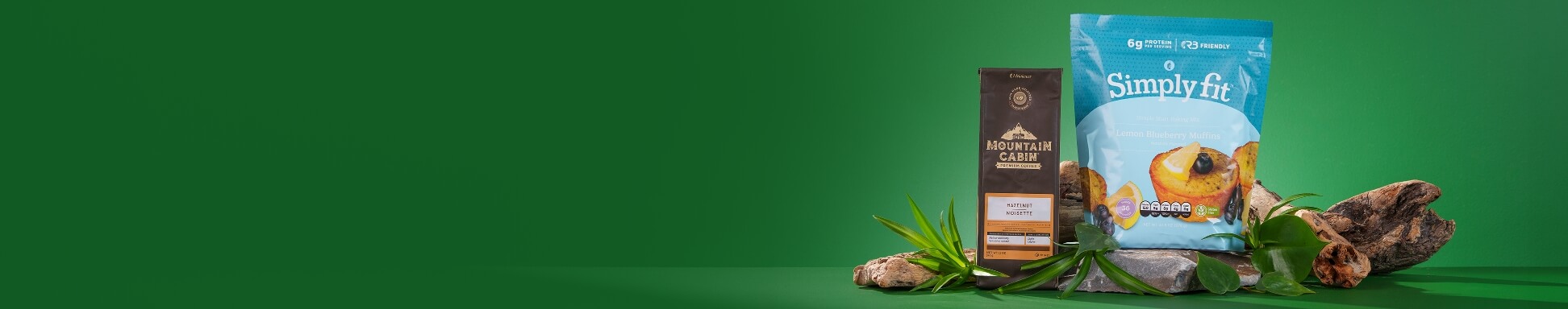 A bag of Mountain Cabin Coffee and a bag of Simply Fit Simple Start Muffin Mix surrounded by natural elements like rocks, leaves, and wood.