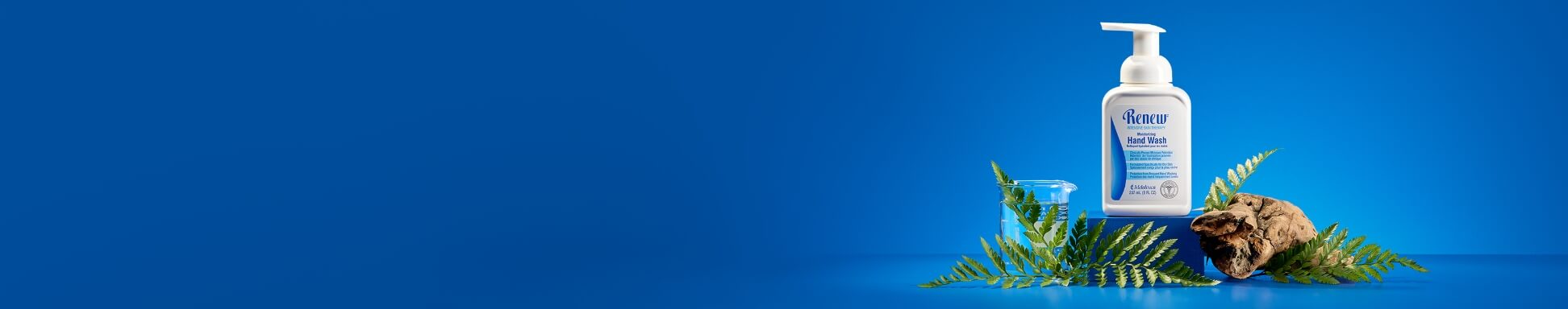 A bottle of hand wash with a pump sits on a blue surface, surrounded by ferns and bark. A clear beaker is nearby, set against a vibrant blue background.