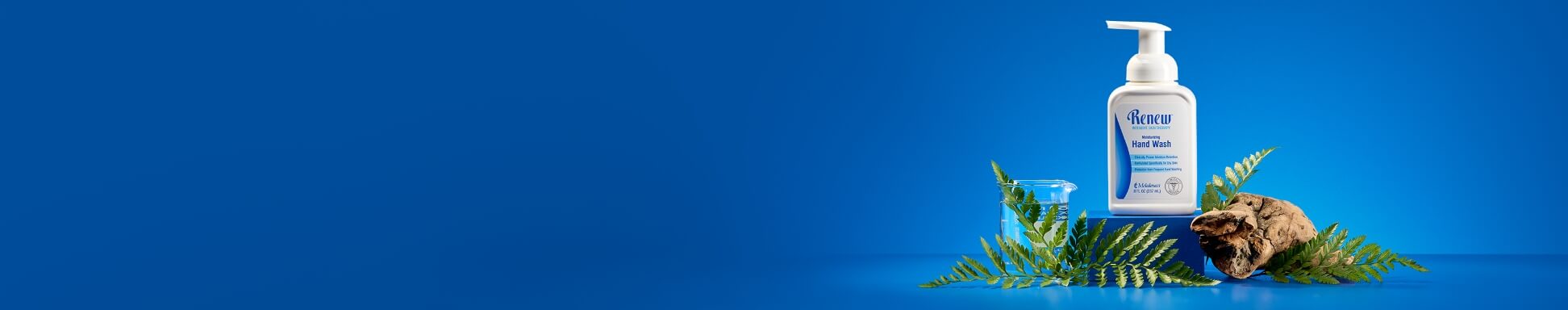 A bottle of hand wash with a pump sits on a blue surface, surrounded by ferns and bark. A clear beaker is nearby, set against a vibrant blue background.