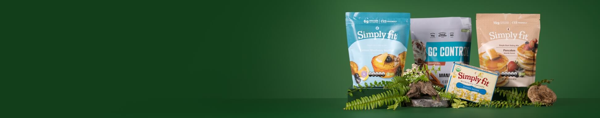 Three Simply Fit food products are arranged on a dark green backdrop with decorative greenery and rocks. The display includes a blue pouch, a gray "GC Control" pouch, a beige pouch, and a matching box placed in front, suggesting a lineup of baking or nutrition products.