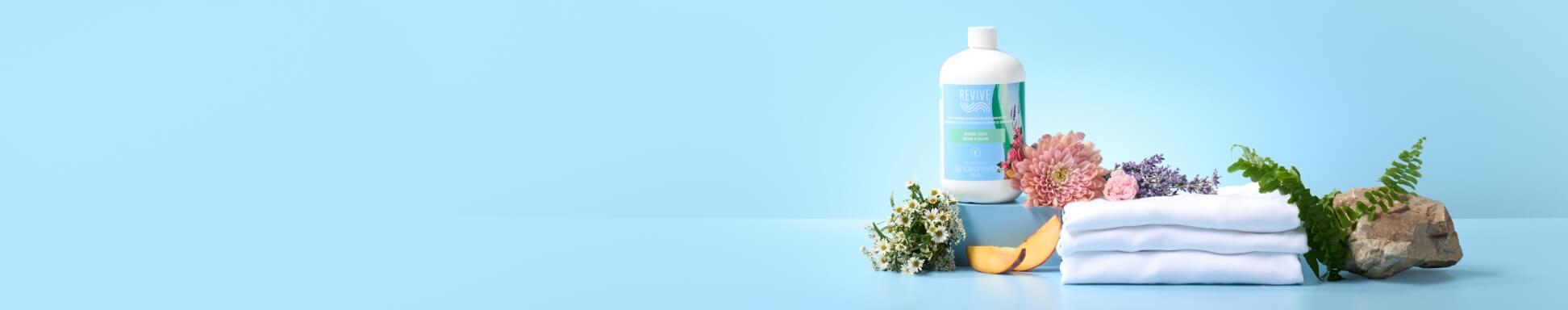 Revive™ Room Spray Refill, pictured with fresh flowers, folded white towels, and a citrus slice on a light blue background.