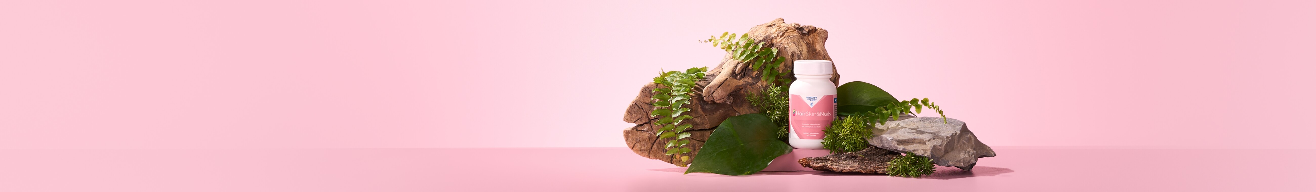 HairSkin&Nails supplement bottle displayed among green leaves, wood, rocks, and moss on a soft pink background, conveying natural beauty.
